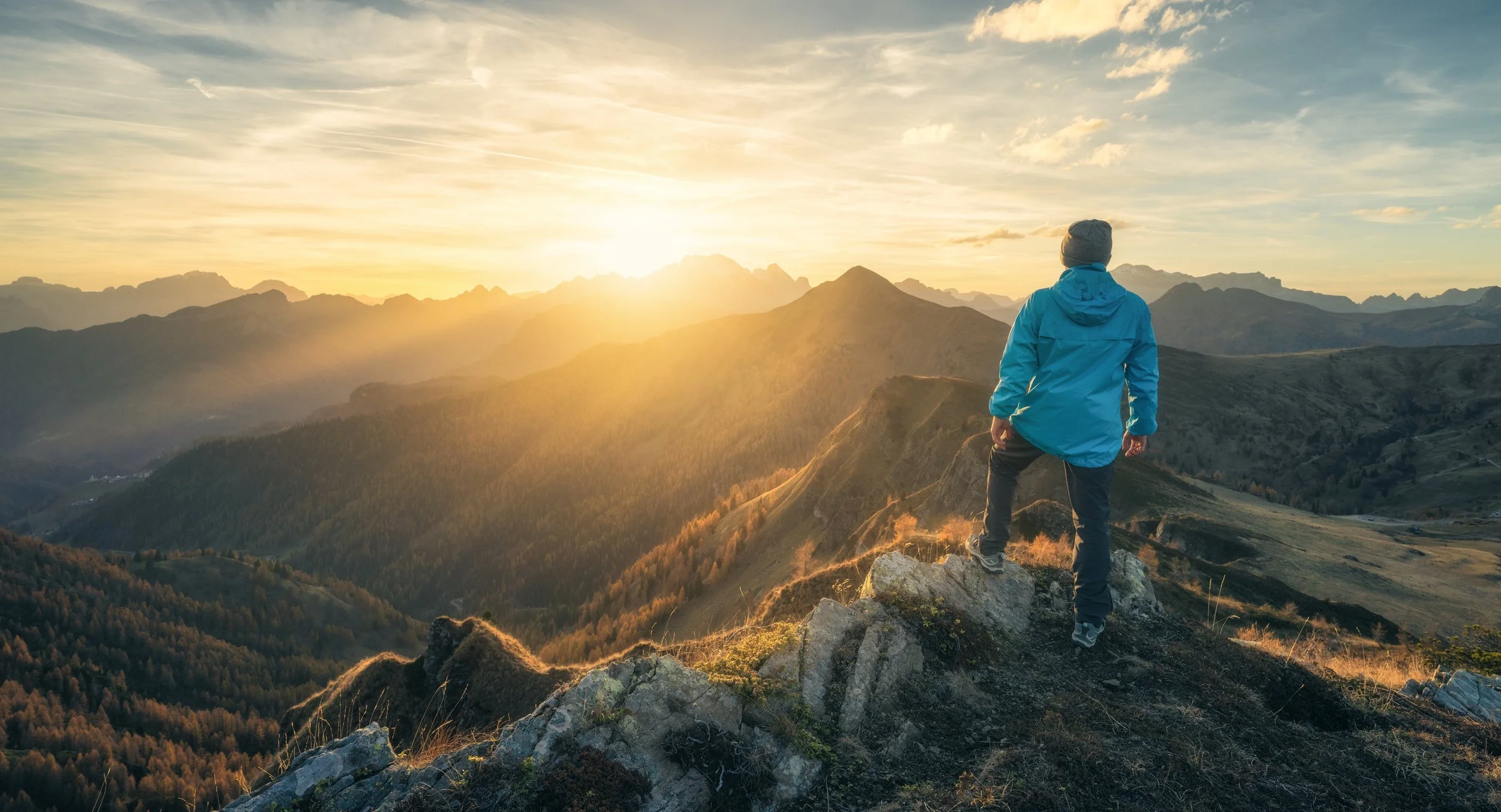 A person in a blue jacket and gray beanie standing on rocks, overlooking a mountain landscape at sunset.