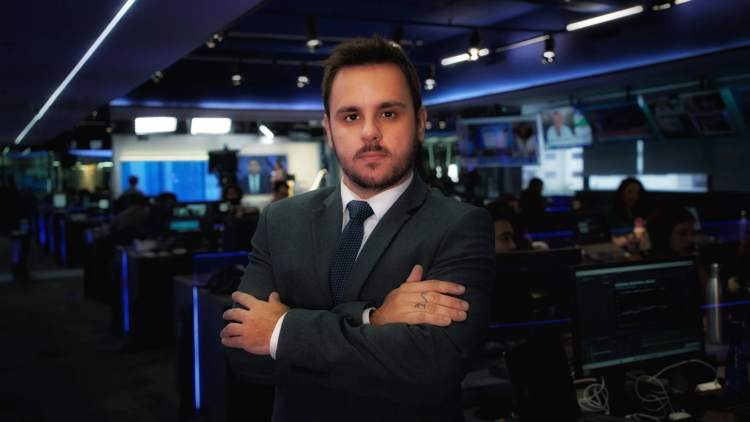 Homem de terno escuro, barba e cabelo curto, com braços cruzados, no ambiente de uma sala de redação ou centro de controle com várias telas e computadores ao fundo.