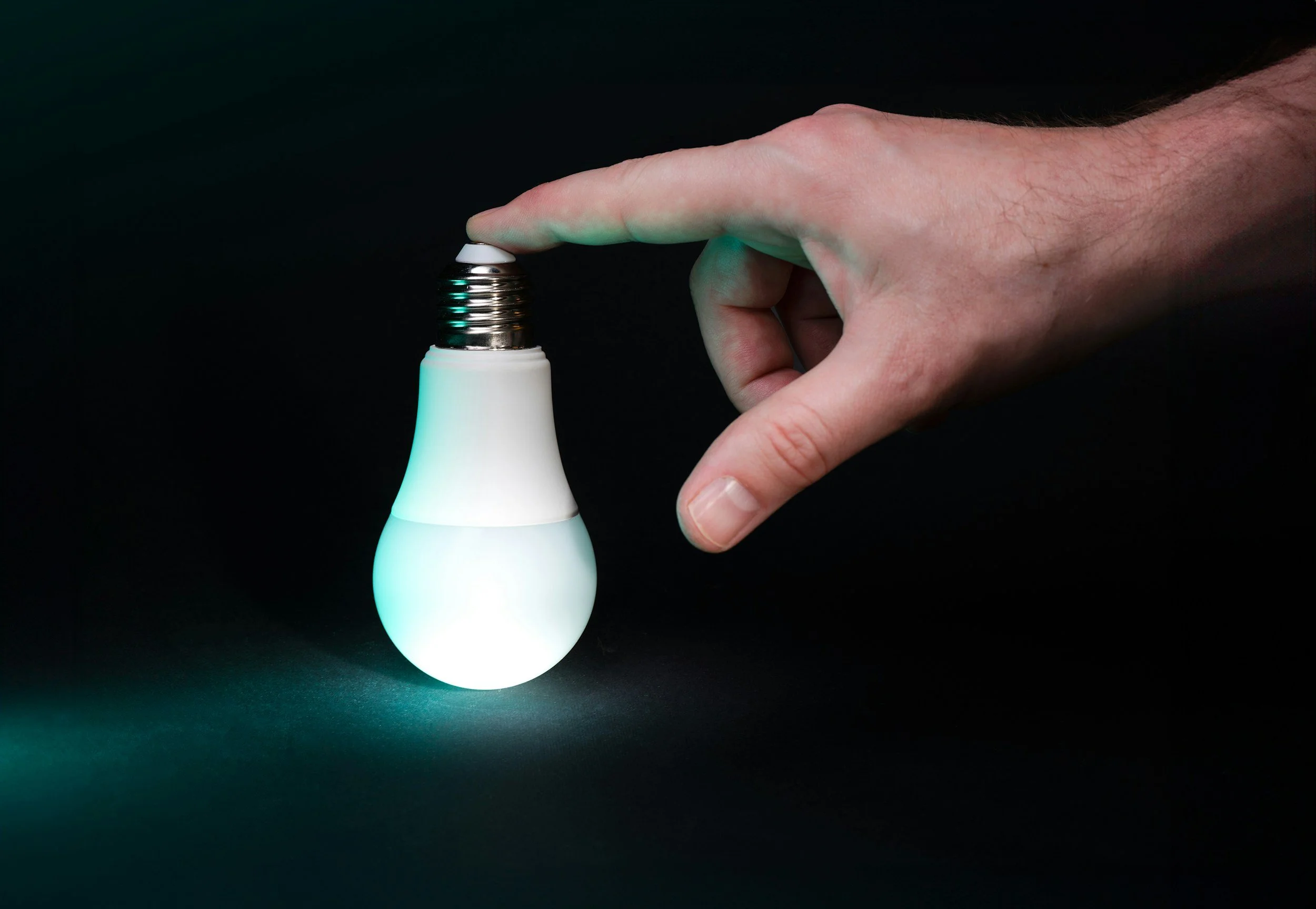 A hand touching the metal base of a glowing LED light bulb against a black background.