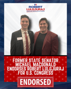 Two men standing together in front of campaign signs. The sign reads, 'Former State Senator Michael Macdonald Endorses Robert Lulgjuraj for U.S. Congress.' Both men are smiling and dressed in business attire.