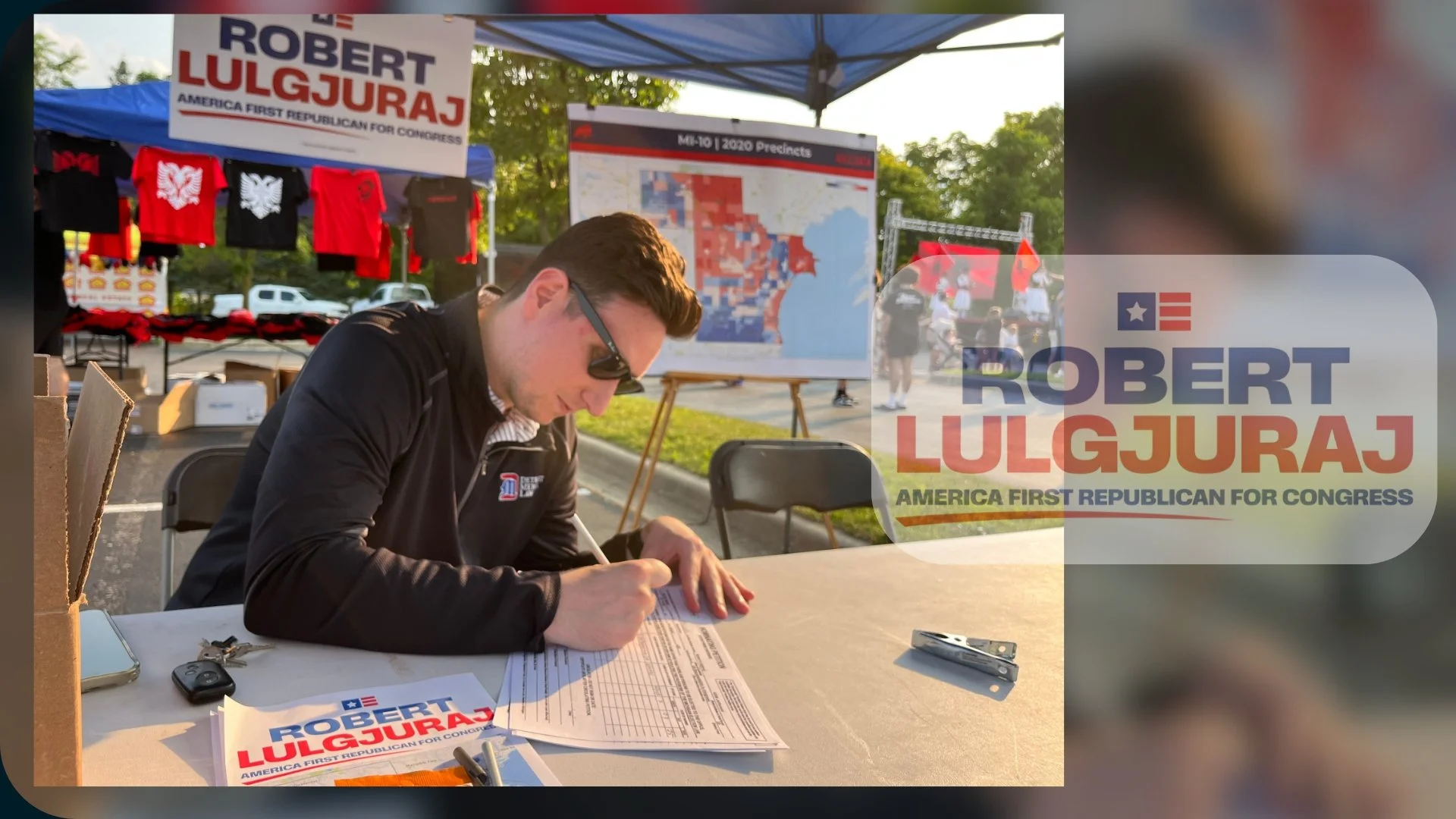 A man wearing sunglasses and a black jacket is sitting at a table outdoors, signing paperwork. A political campaign sign for Robert Lulgjuraj, a Republican candidate, is visible on the table and in the background, along with campaign merchandise such