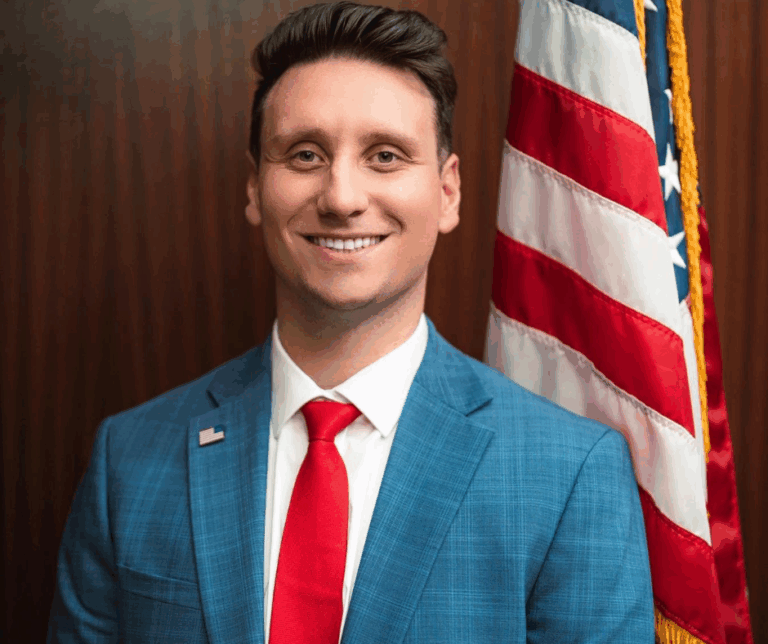 Robert Lulgjuraj, a man in a blue suit and red tie standing in front of an American flag with a wooden background.