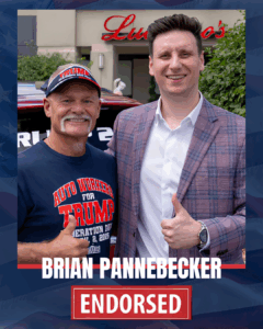 Two men smiling at a public event, one wearing a blue campaign t-shirt and the other in a suit, with a car and a building with a sign in the background.