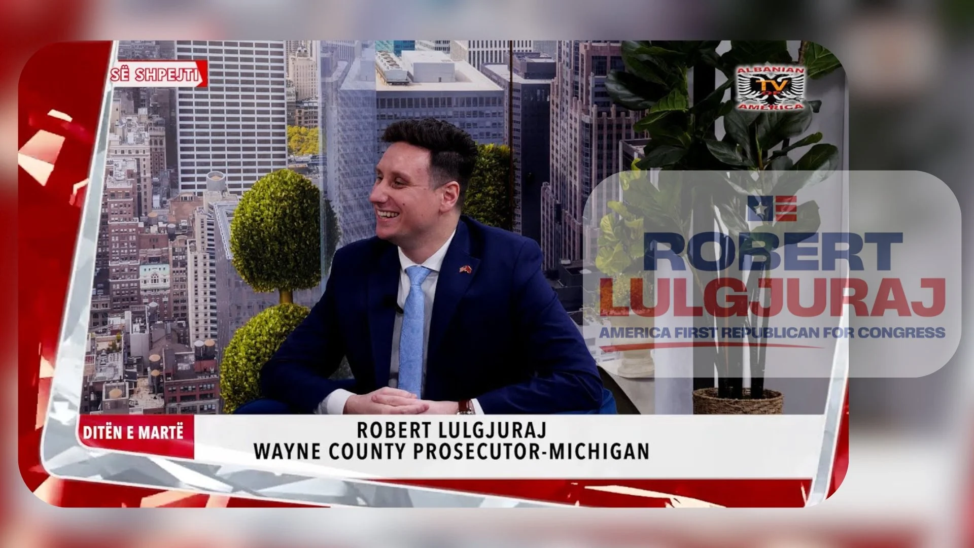 A man in a dark suit, white shirt, and light blue tie sitting at a desk or table, smiling in front of a cityscape background with tall buildings and some greenery, during a television interview on Albanian TV America. The screen displays his name, Ro