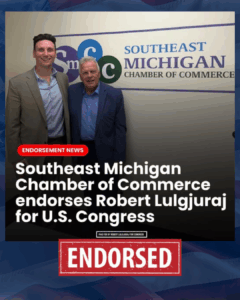 Two men standing in front of a sign that reads Southeastern Michigan Chamber of Commerce, with one man in a dark blazer and white shirt, and the other in a blue blazer, both smiling.