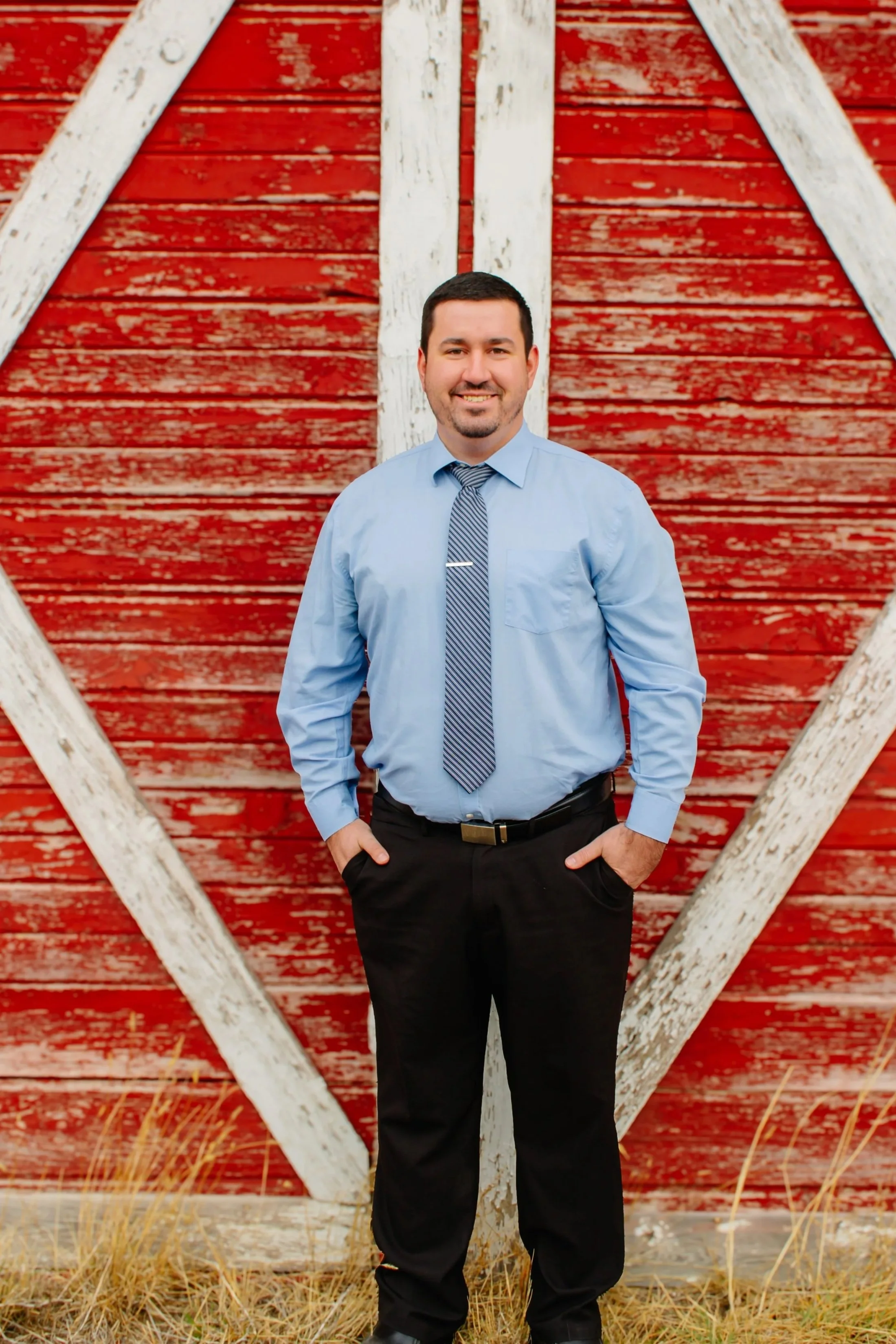 A man with short dark hair and a beard, wearing a light-colored striped button-up shirt with a blue tie, standing outdoors against a wooden barn paneled wall, smiling.