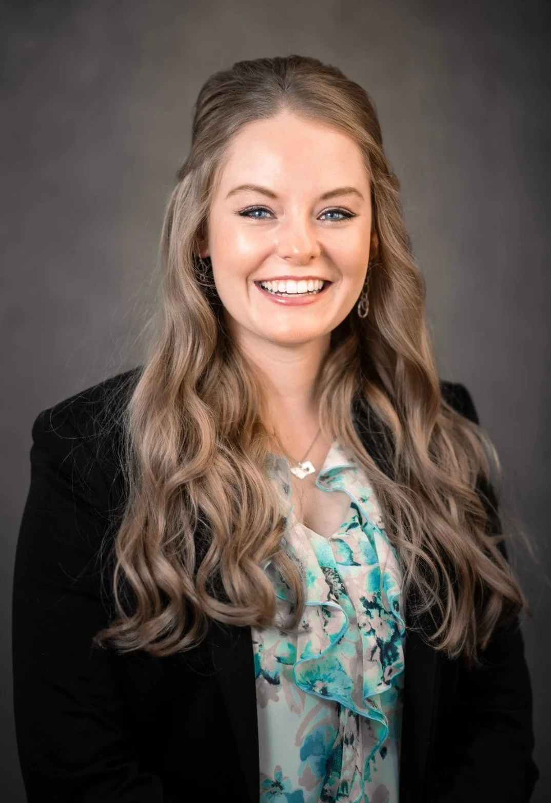 A woman with long, wavy blonde hair, wearing earrings, a patterned blouse, and a black blazer, smiling against a gray background.