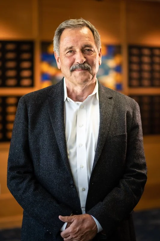 A middle-aged man with gray hair and a mustache, wearing a dark blazer and white shirt, standing in a room with wood paneling and colorful artwork in the background.