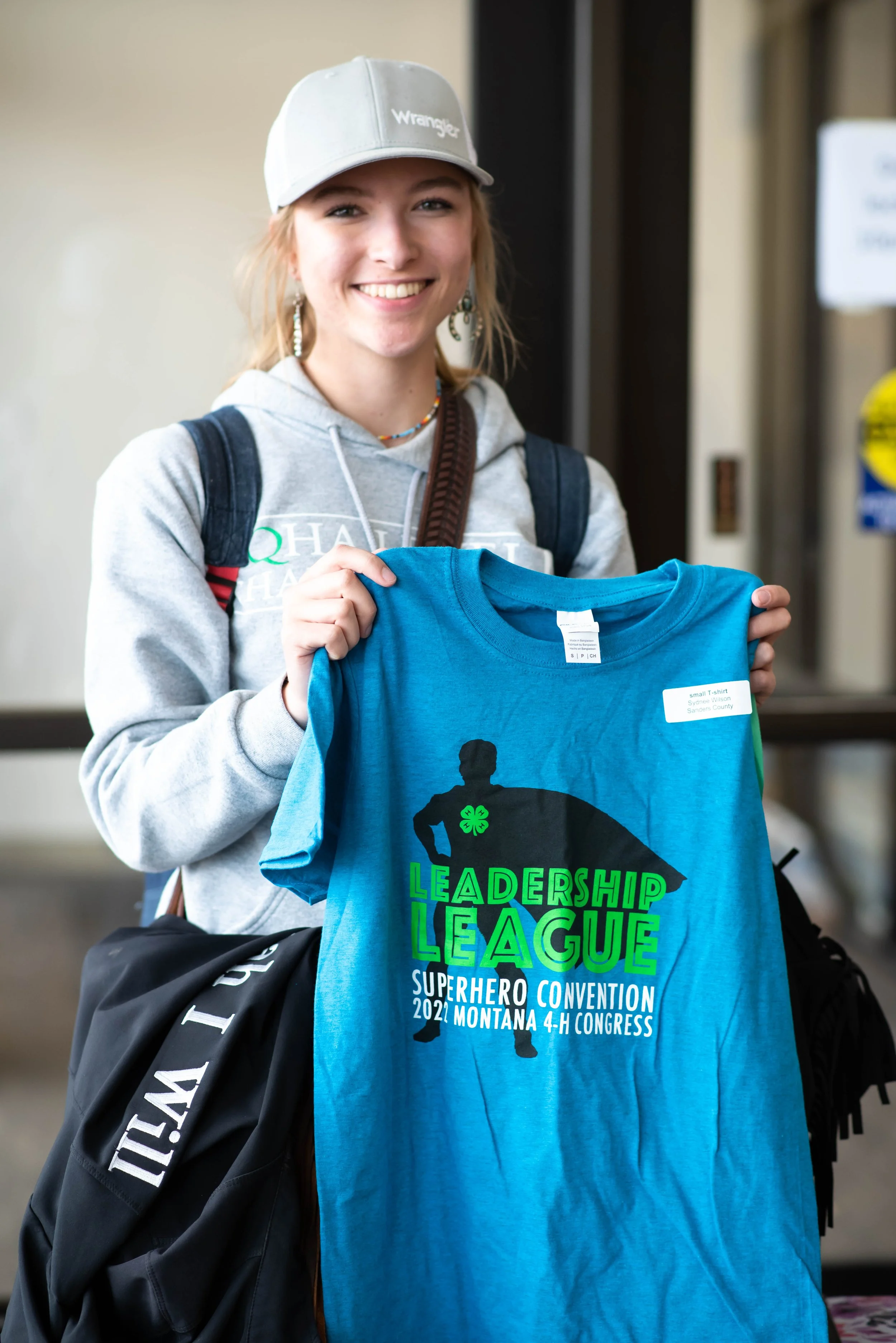 A young woman with blonde hair, smiling, holding a blue T-shirt with a superhero graphic and text promoting Leadership League at a 4-H Congress in Montana, wearing a gray hoodie, a gray cap, and a backpack.