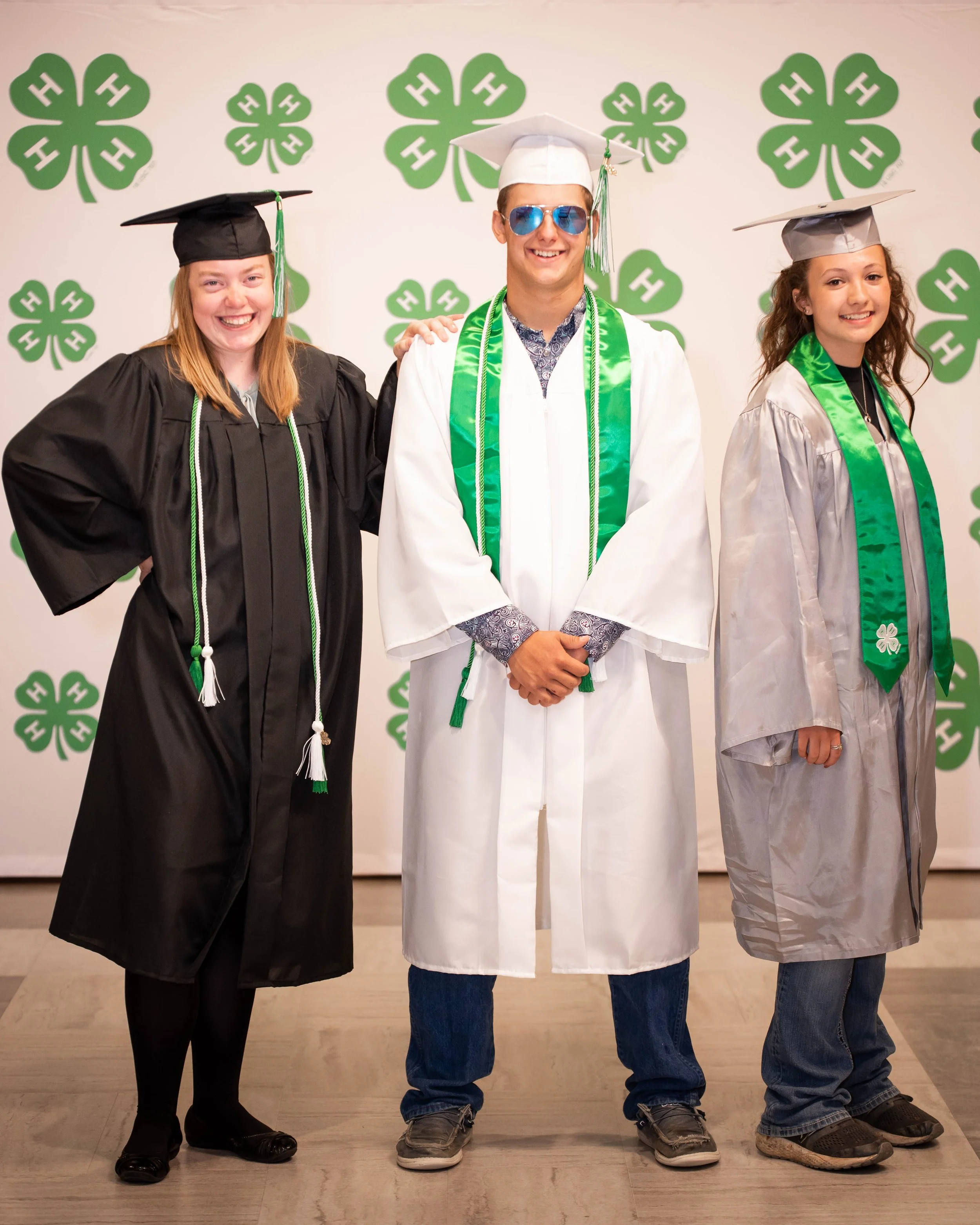 Three people in graduation caps and gowns smiling for a photo, standing against a backdrop decorated with green four-leaf clover patterns.