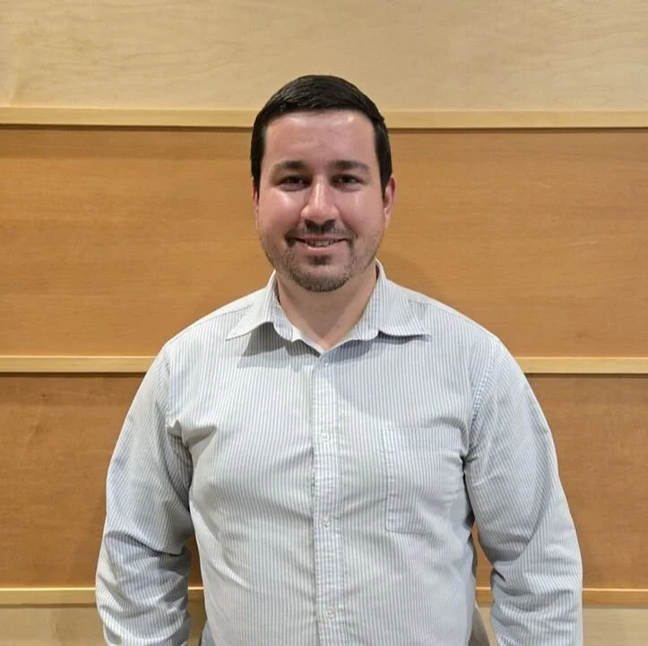 A man with short dark hair and a beard, wearing a light-colored striped button-up shirt, standing indoors against a wooden paneled wall, smiling.