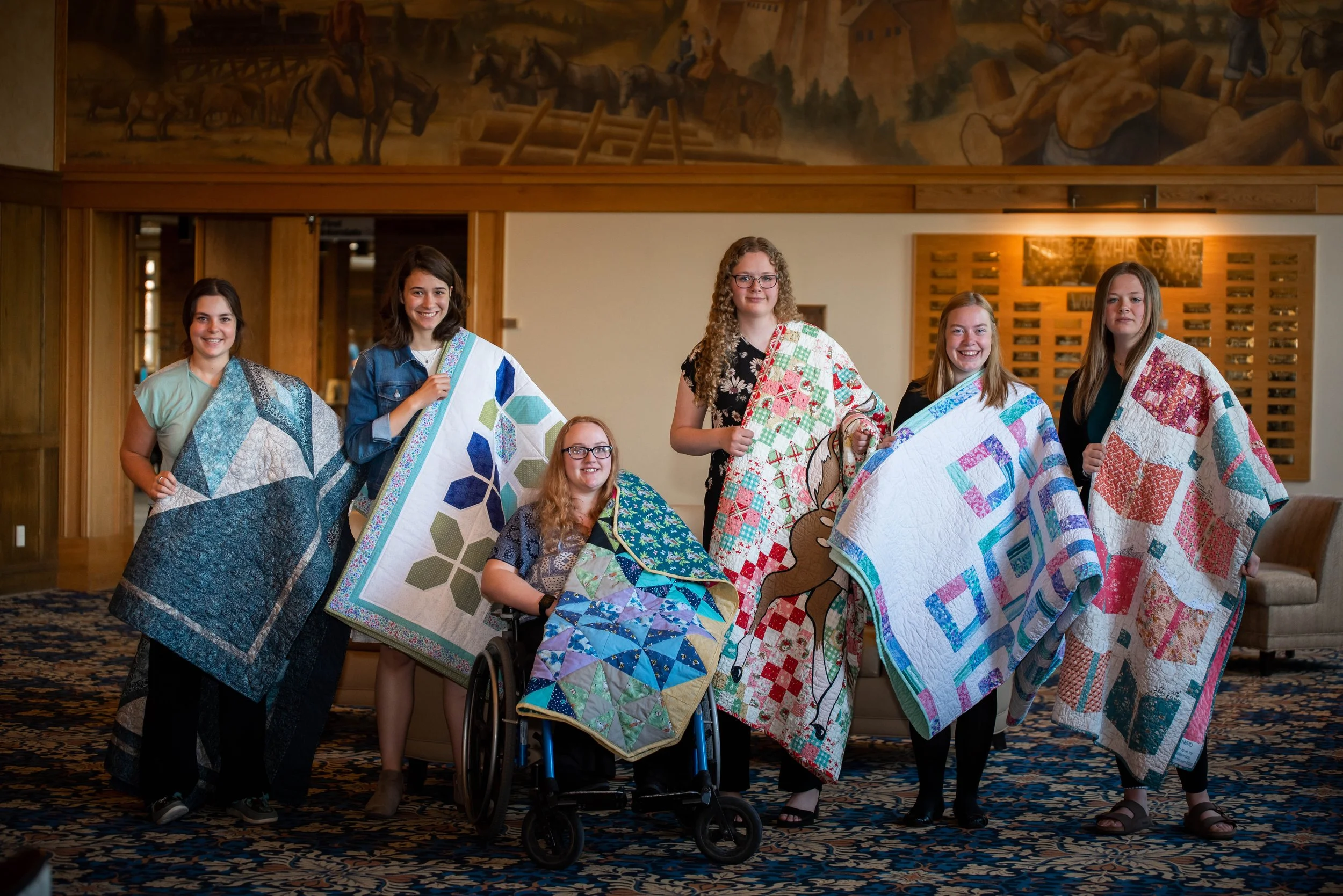 Six young women displaying handmade quilts indoors, with wooden paneling and mural artwork in the background.