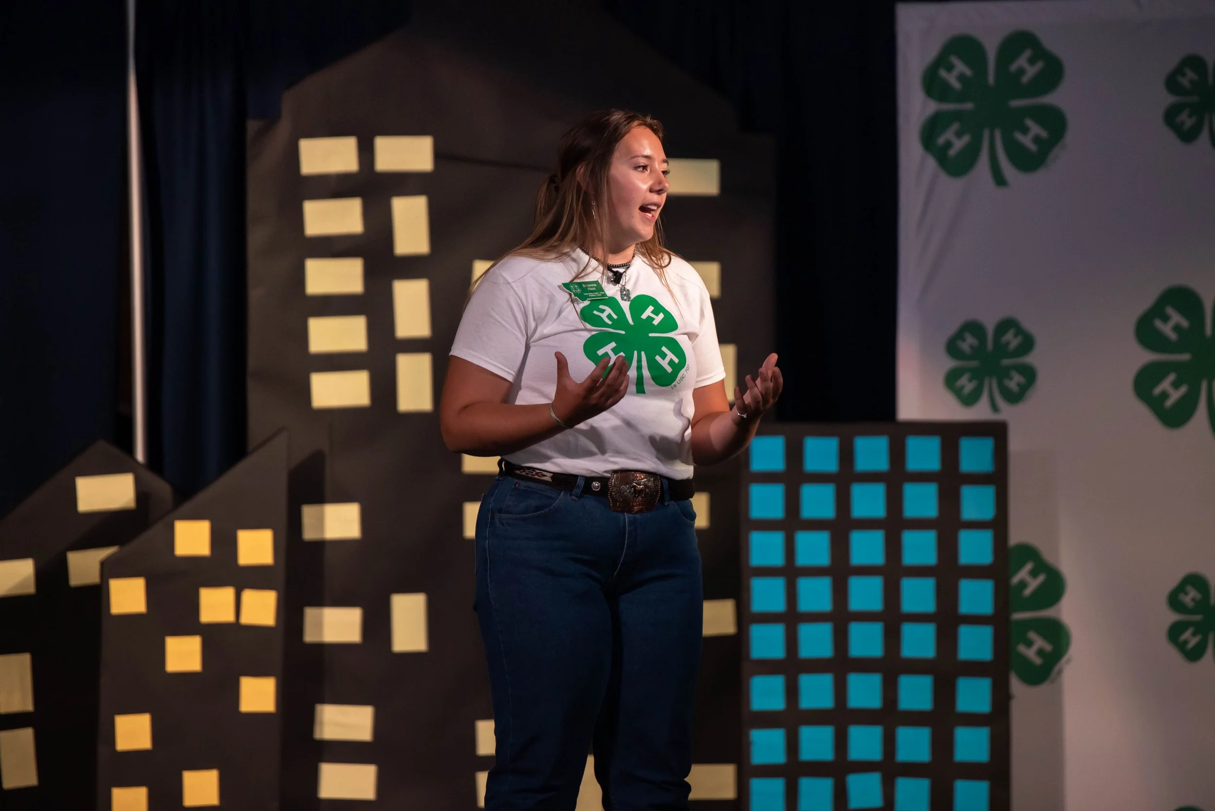 A young woman giving a presentation at a conference, standing in front of a backdrop with green hashtags and cityscape illustrations, wearing a white shirt with the four-leaf clover and an 'H' logo.