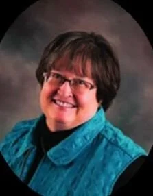 A smiling woman with short brown hair and glasses, wearing a blue jacket and black shirt, posing for a portrait against a dark, blurred background.
