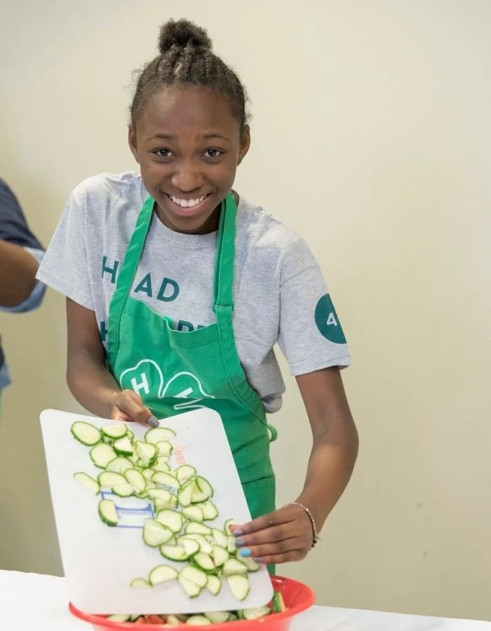 Smiling girl in gray T-shirt and green apron chopping cucumbers over a red bowl.