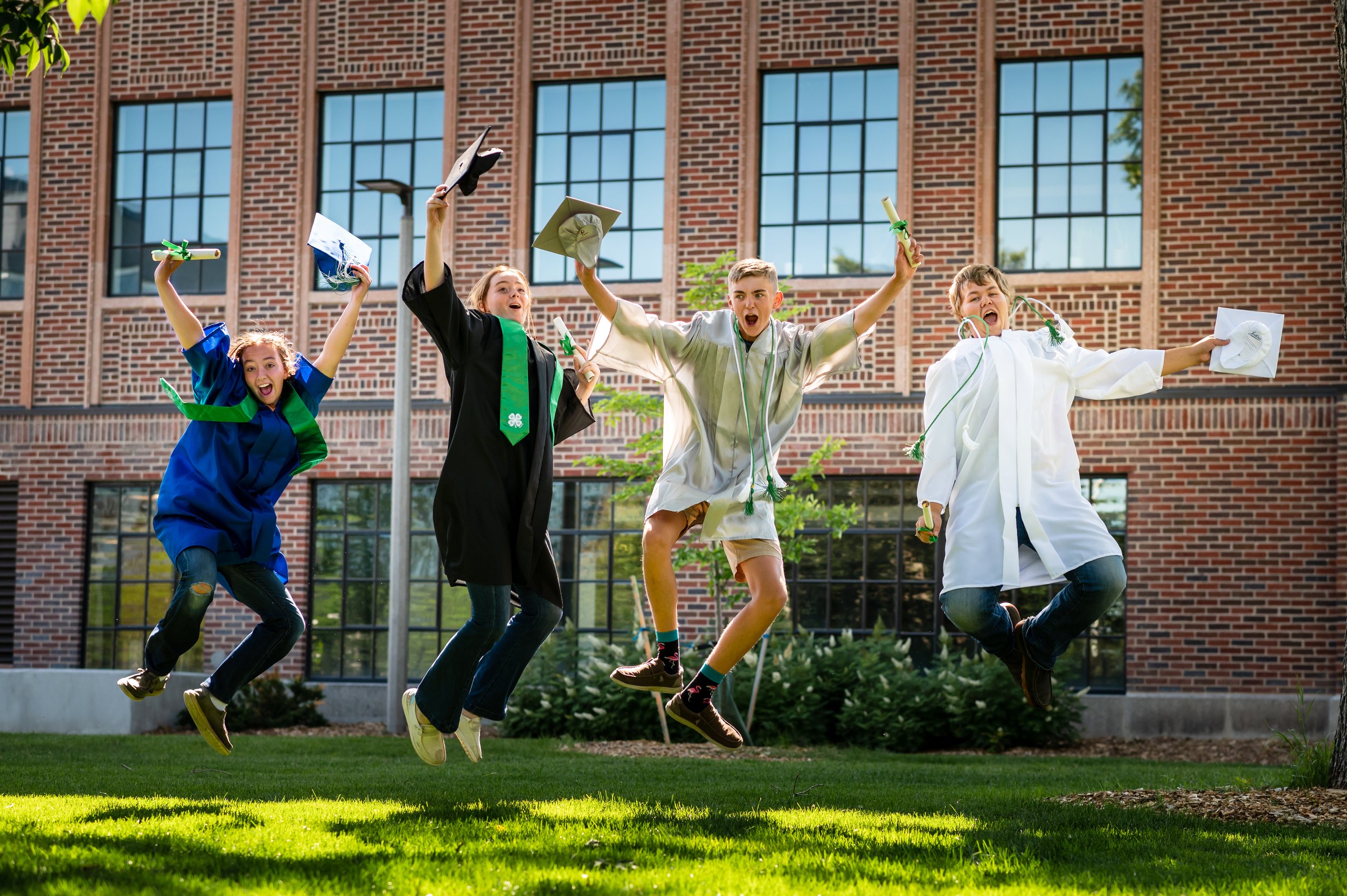 Four students in caps and gowns jumping in the air after graduation, outdoors in front of a brick building.