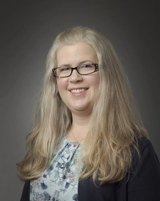 A woman with long, blonde hair wearing glasses, smiling, dressed in a floral blouse and a dark blazer,, against a dark grey background.