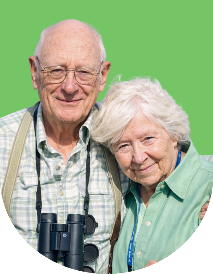 An elderly man and woman smiling, with green screen background, the man wearing glasses, plaid shirt, binoculars around his neck, and the woman in a green shirt with a blue lanyard.