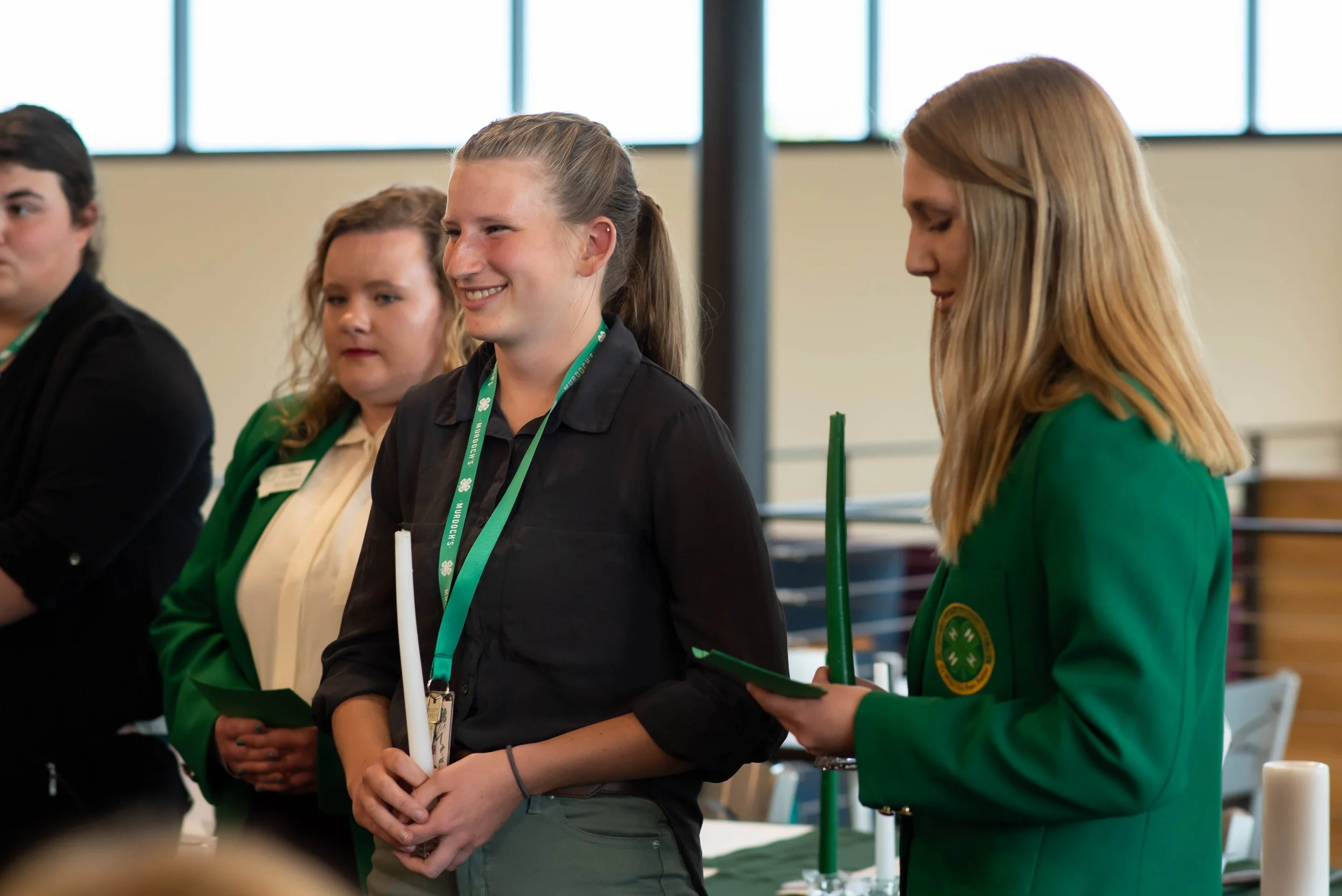 Group of young women at an indoor event, with one woman in a green jacket holding a candle, and another woman in a green blazer reading from a paper, smiling.