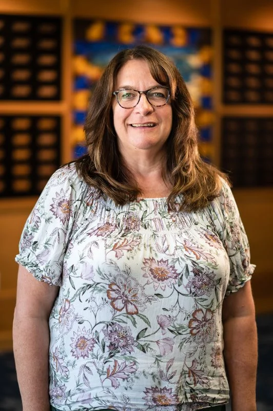 A smiling woman with glasses in a library or bookstore, wearing a floral blouse.