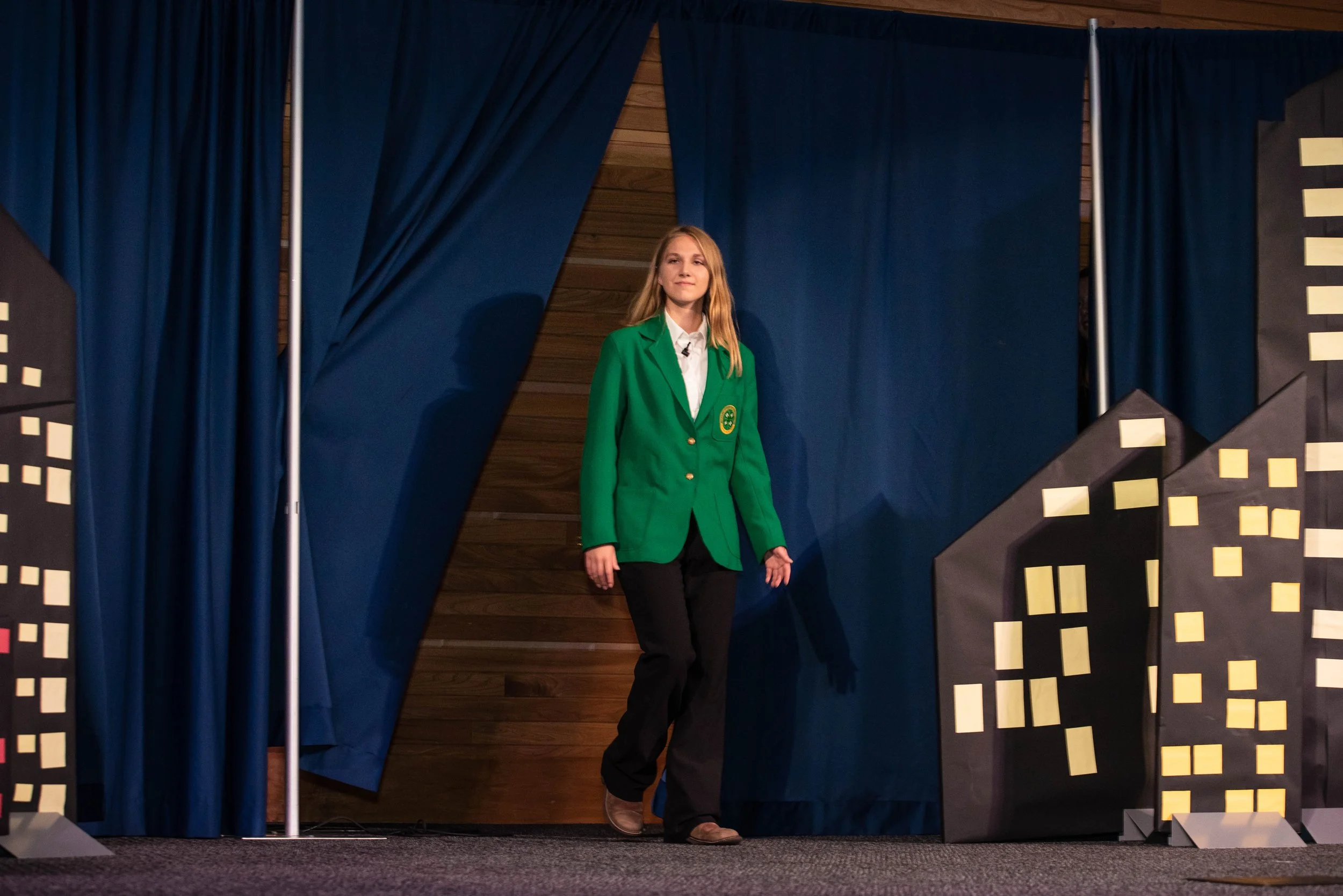 Young woman in a green blazer walking onto a stage with cityscape cutouts and blue curtains.
