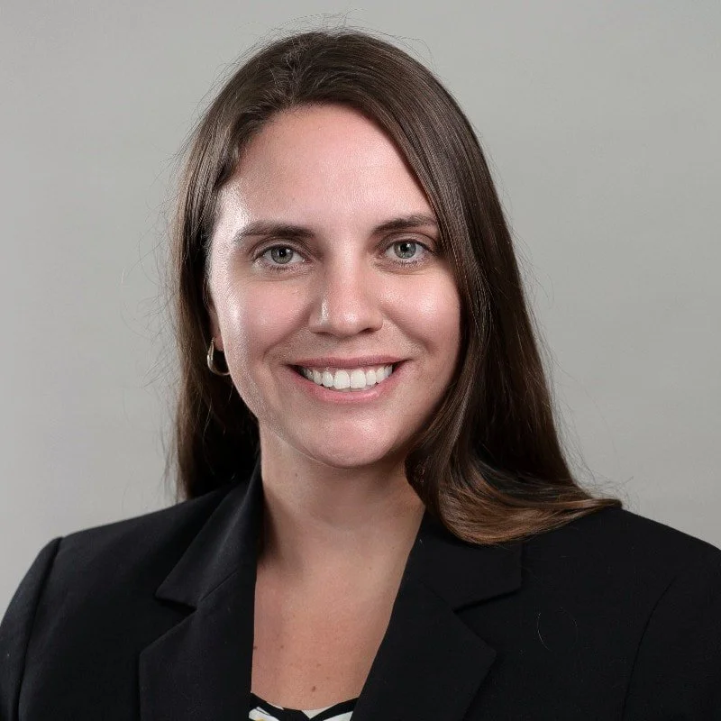 A professional woman with shoulder-length brown hair, wearing a black blazer and a patterned top, smiling against a plain gray background.