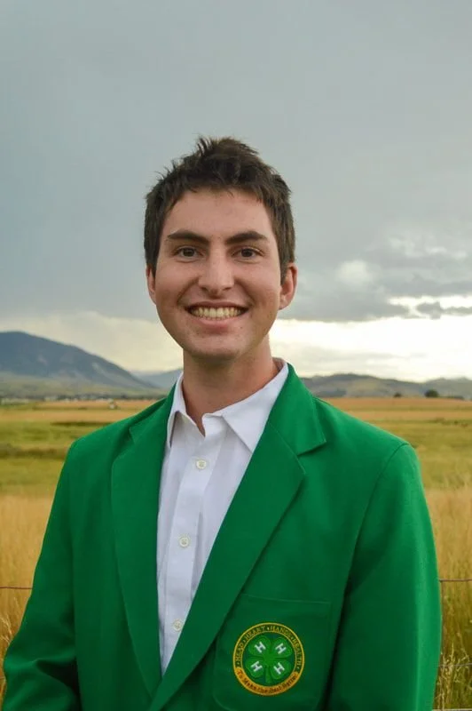 Young man in a green jacket with a university emblem, standing outdoors in a grassy field with mountains and cloudy sky in the background.