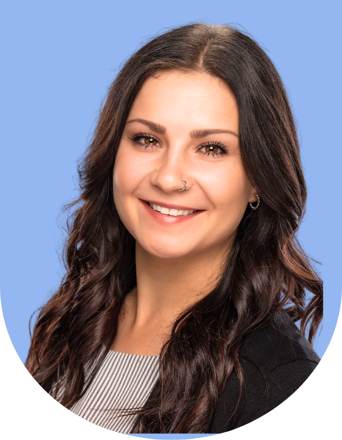 Portrait of a young woman with long dark wavy hair, wearing a nose ring, earrings, a striped shirt, and a black blazer, smiling against a blue background.