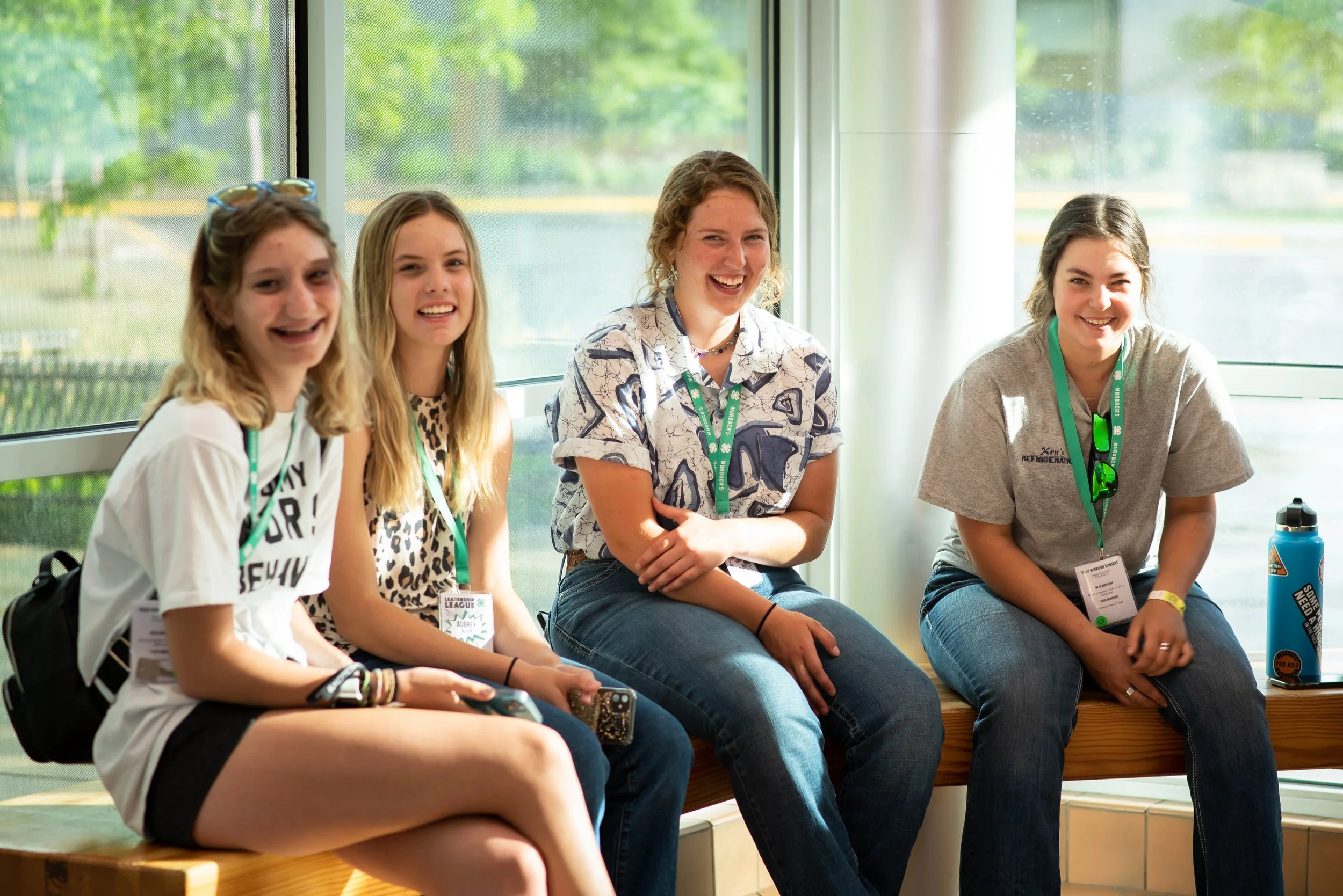 Four teenage girls sitting on a wooden bench near large windows, smiling and enjoying each other's company during daytime, with green trees visible outside.