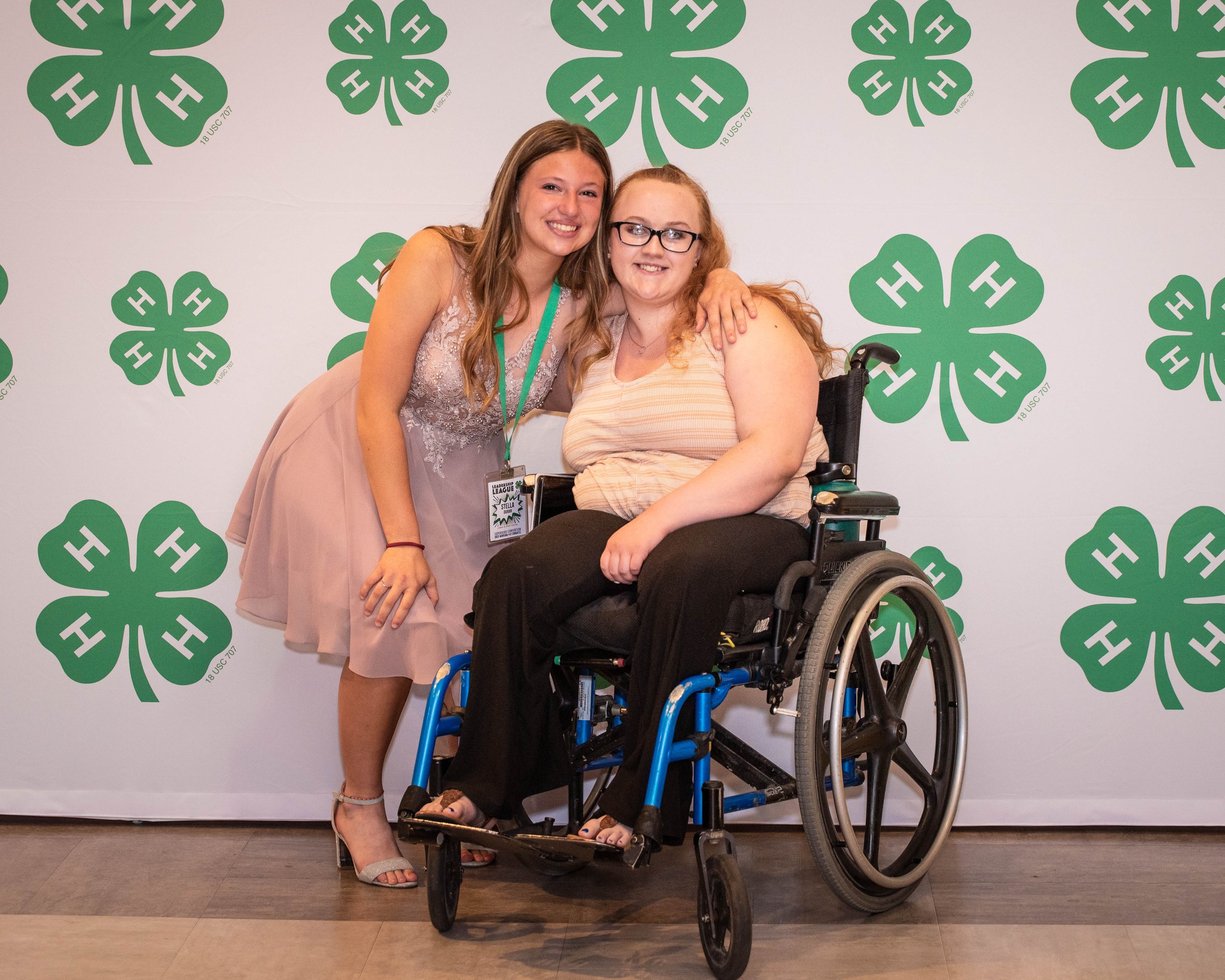 Two women, one standing and one in a wheelchair, smiling and posing together in front of a backdrop with green four-leaf clovers.