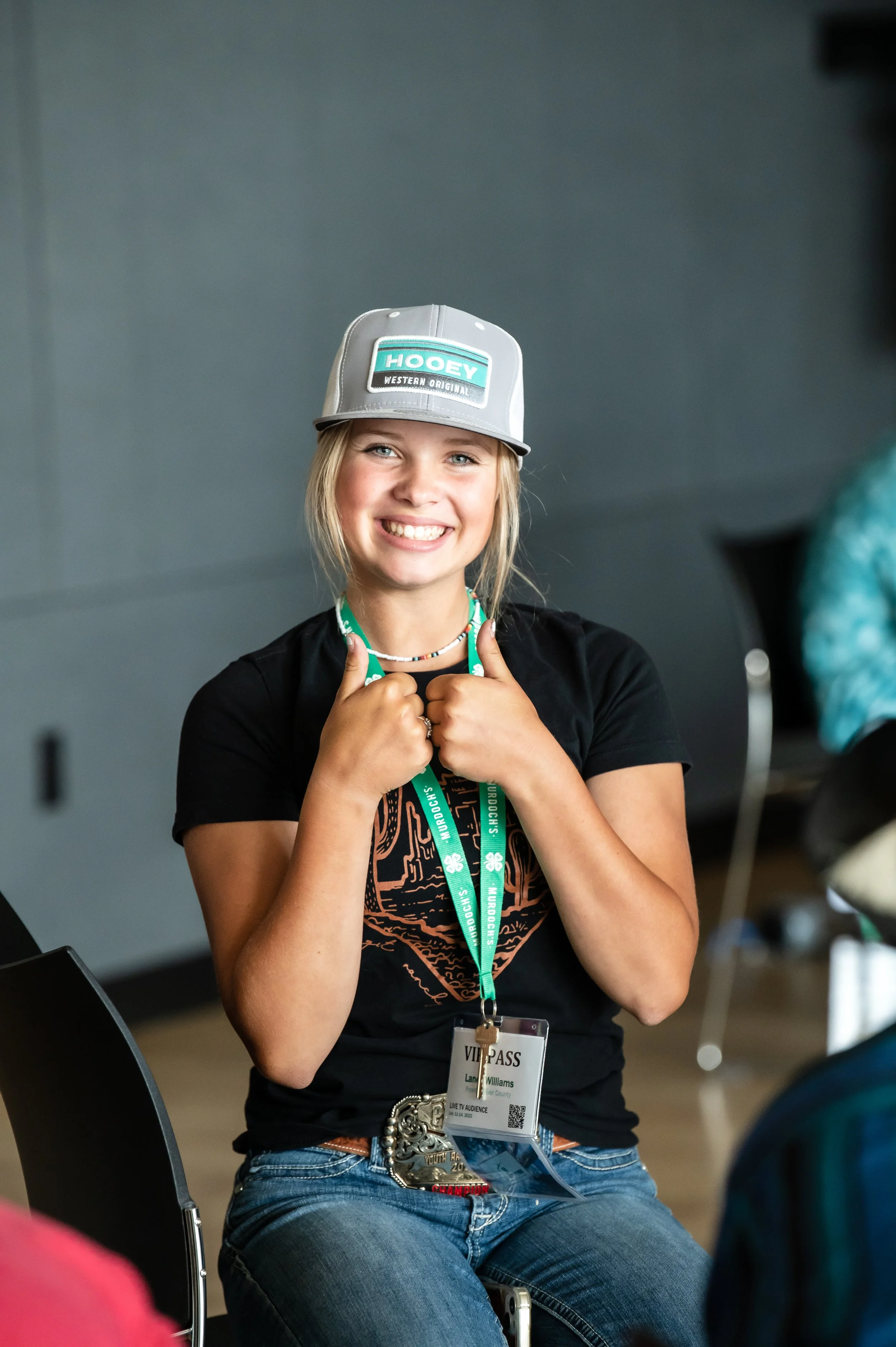 A young woman with blonde hair, smiling and giving two thumbs up, wearing a gray cap with a logo, a black T-shirt, a VIP pass lanyard, and jeans, sitting in a chair indoors.