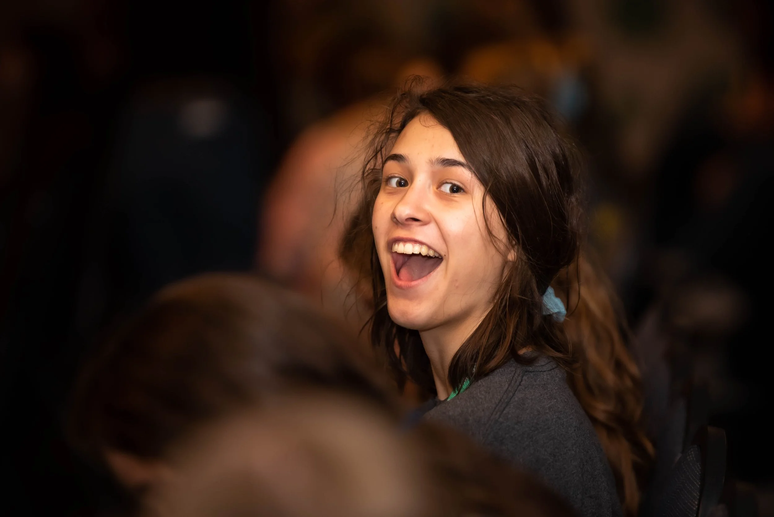A young woman with brown hair smiling and looking back in a crowded indoor setting.