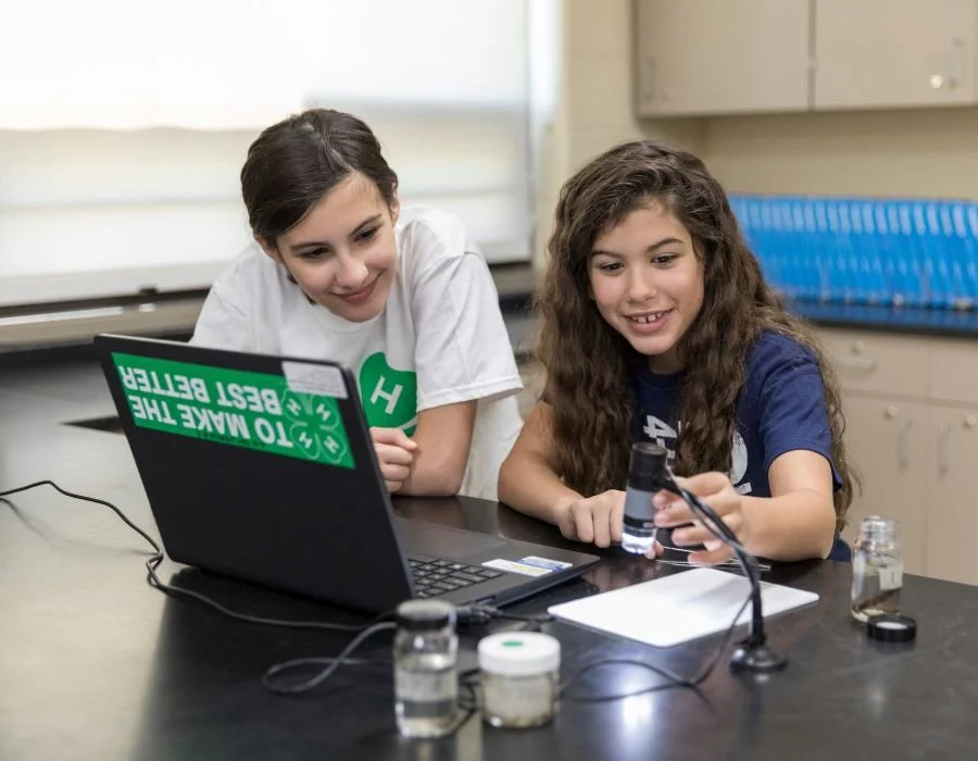 Two young girls conducting science experiments with a laptop and lab equipment at a classroom counter.