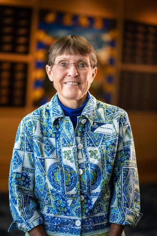Smiling woman with short gray hair and glasses, wearing a blue and white patterned jacket, standing indoors with a blurred background of a lobby or office reception area.