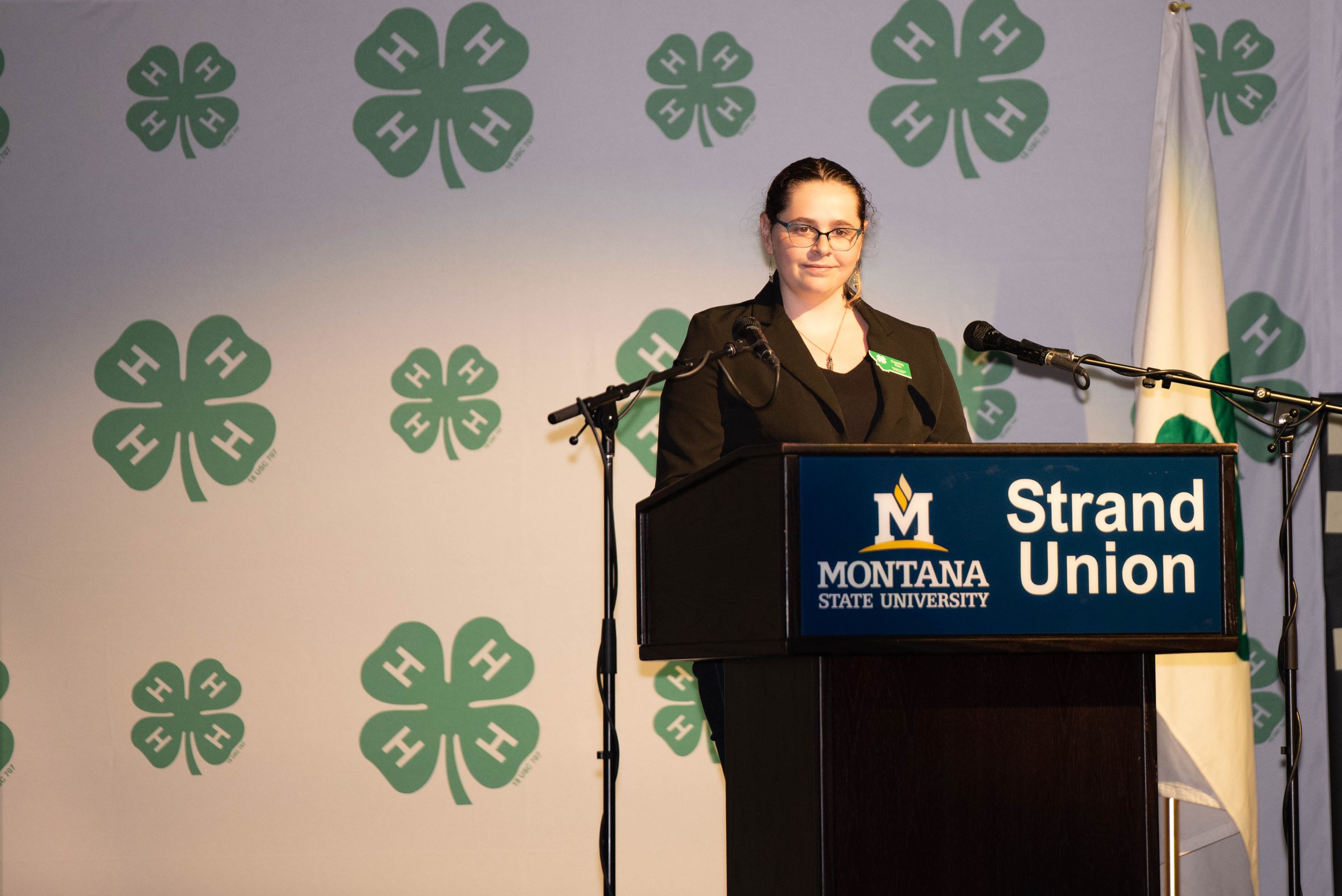 A woman standing at a podium with a sign that reads 'Montana State University Strand Union,' speaking or preparing to speak. Behind her, there is a backdrop with green four-leaf clover symbols and a white flag on the right side.