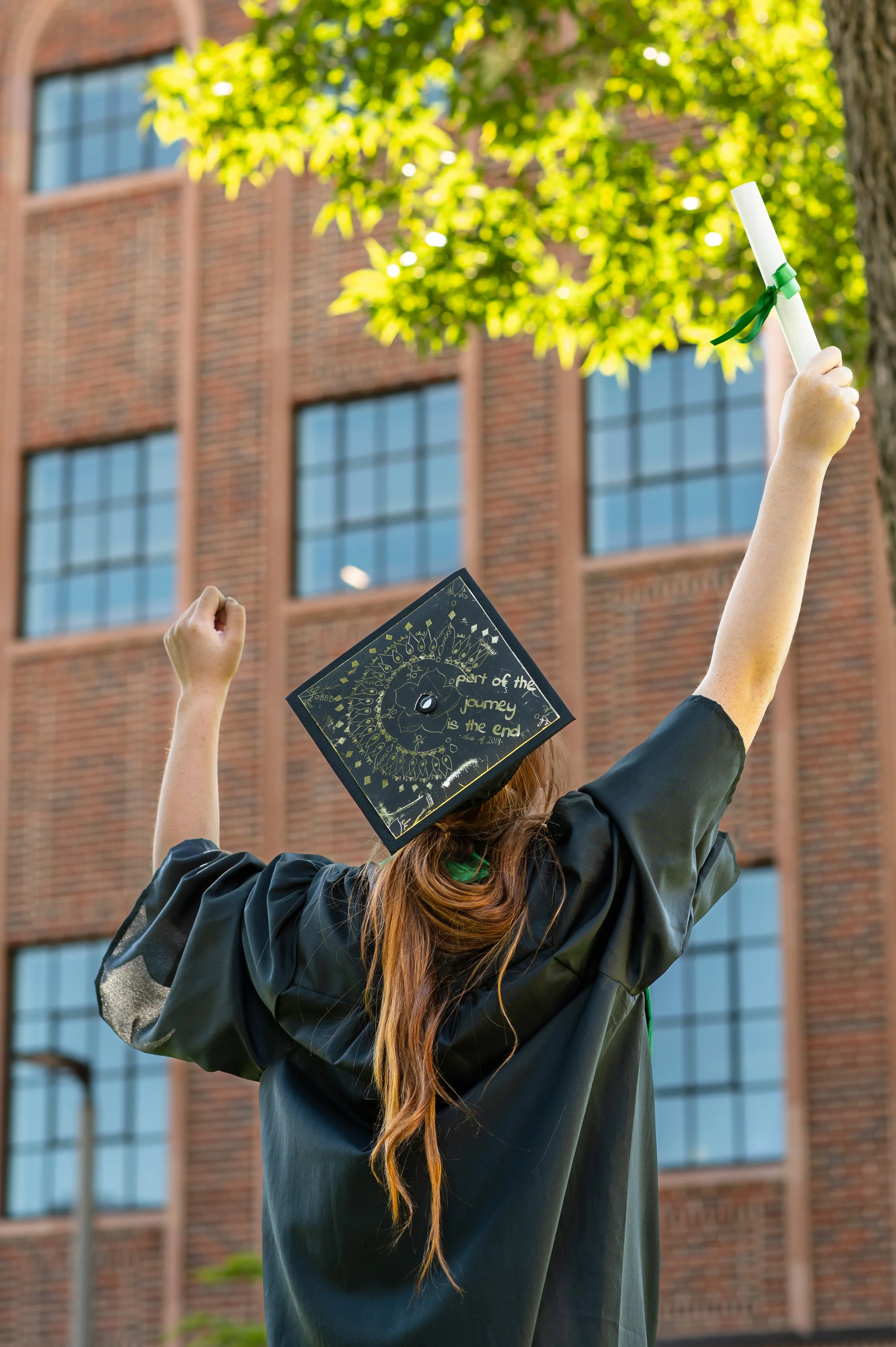 Graduate celebrating outdoors in cap and gown with diploma raised, with a brick building and green trees in the background.