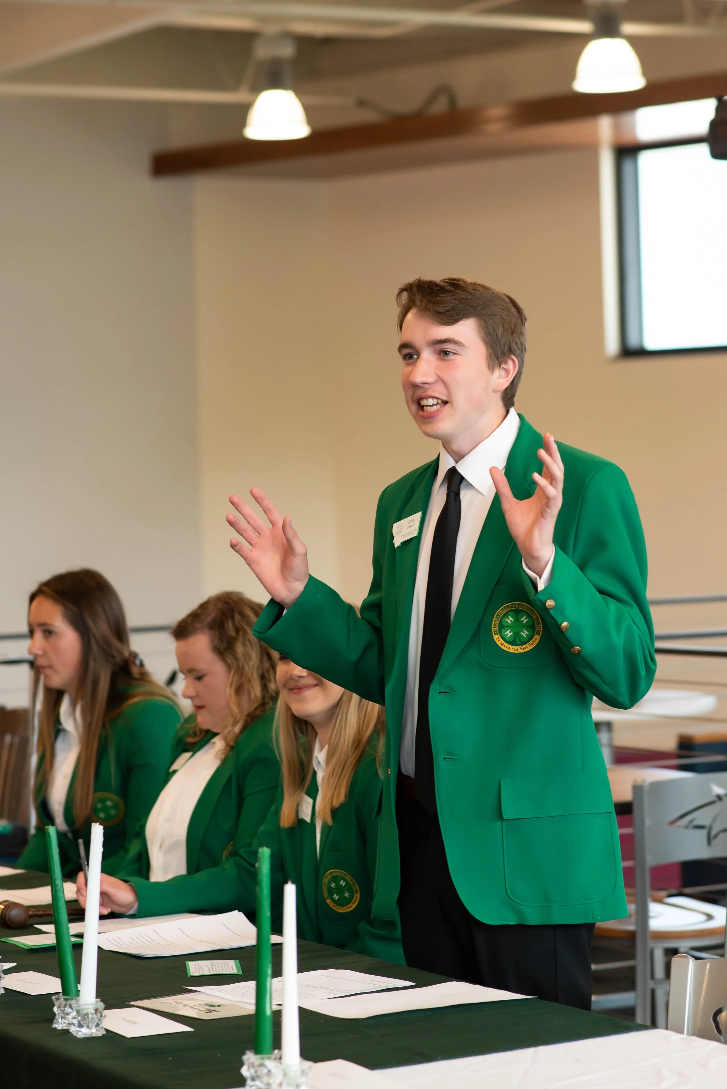 A young man in a green blazer with a circular emblem on the pocket, white shirt, and black tie, appears to be speaking or presenting to an audience. He is standing behind a table with papers and candles, next to three young women also in green blazers, sitting at the table.