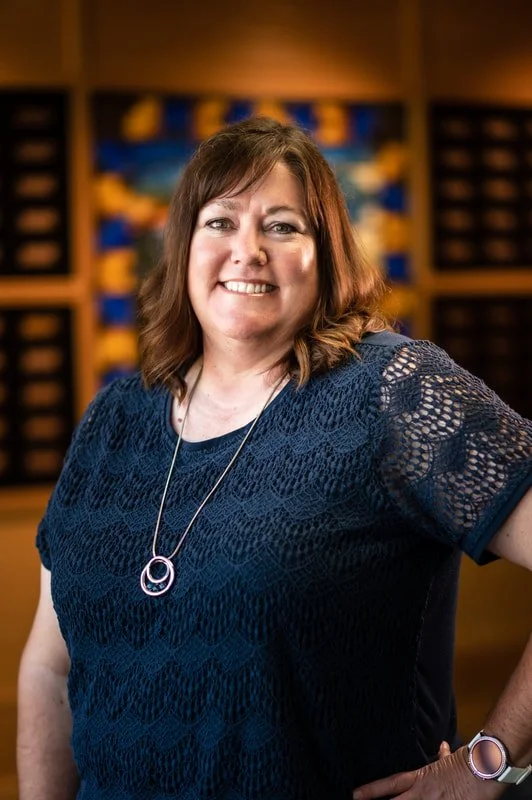 A woman with brown hair smiling, wearing a navy lace top and a long necklace, standing in front of a blurred background of a bookshelf or storage unit.