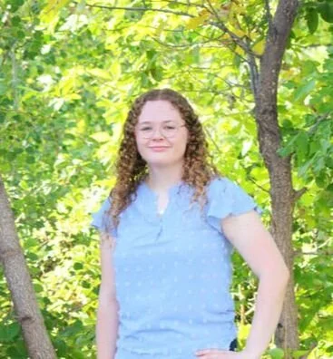 Young woman with curly hair wearing a light blue dress standing outdoors near green trees on a sunny day.