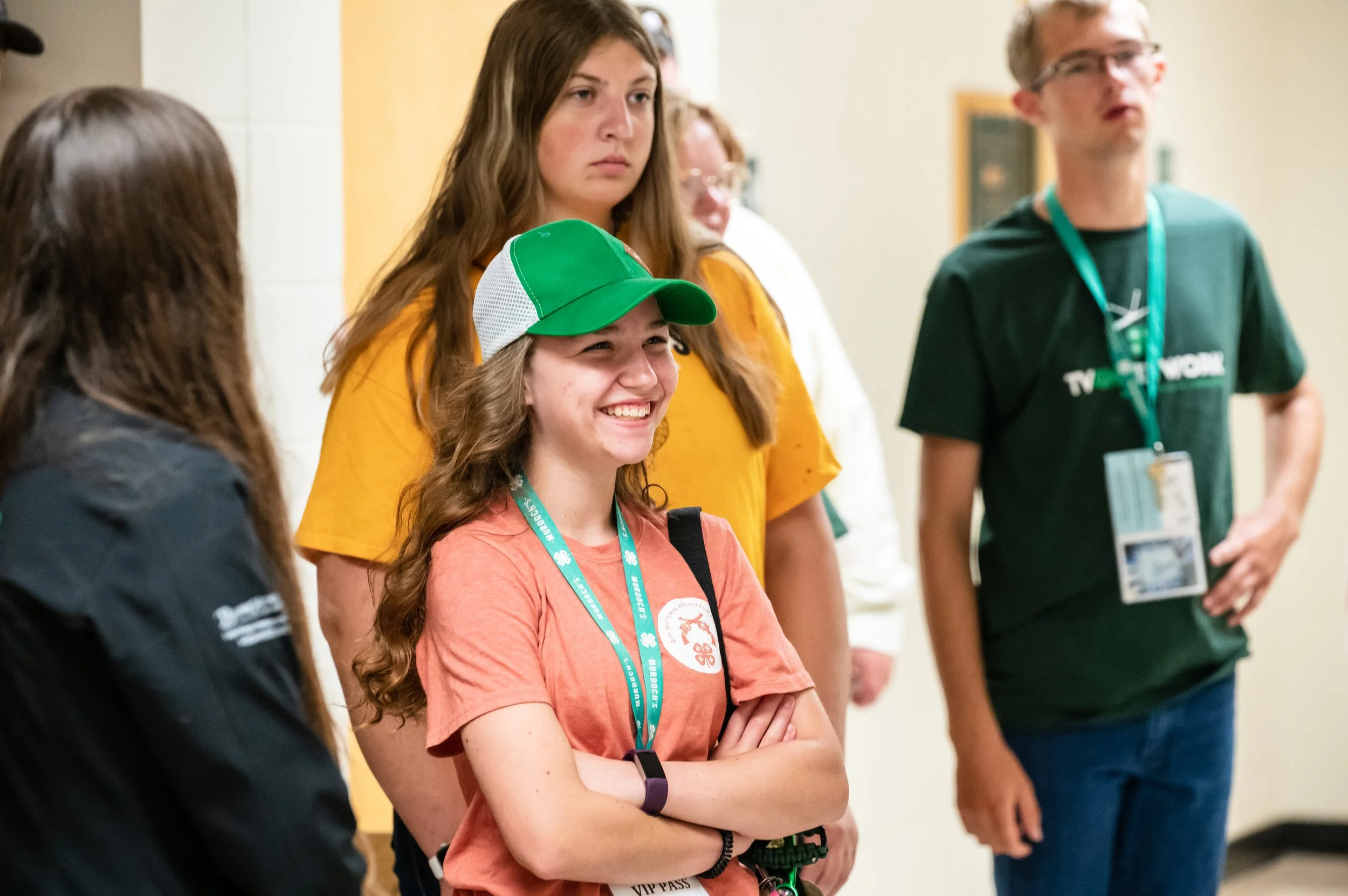 A group of young people indoors, with three girls and one man, standing near a wall. The girl in the front is smiling, wearing a green and white baseball cap, an orange t-shirt, and has a VIP pass hanging from a lanyard.