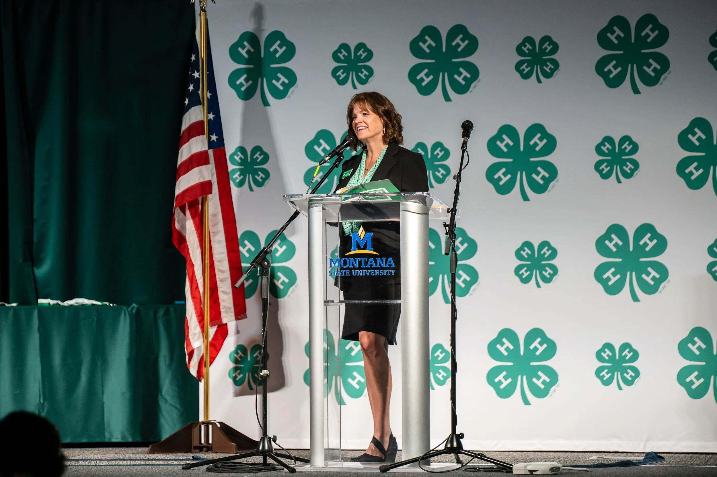 A woman standing at a podium, speaking at an event for Montana State University, with an American flag next to her and a backdrop decorated with Montana State University 4-H logos.