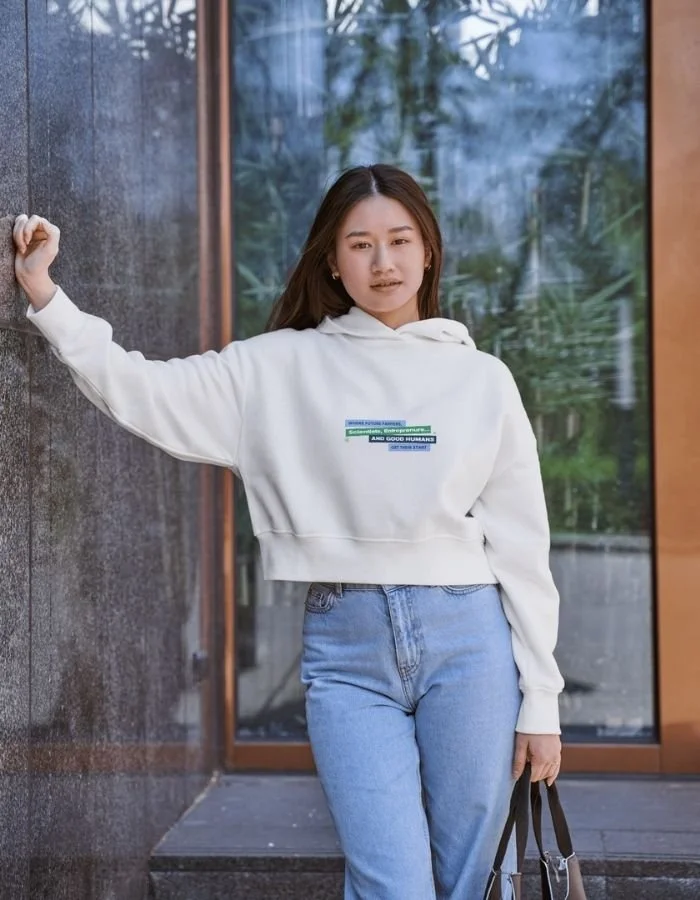 Young woman standing outdoors in front of a glass door, leaning against a wall with one arm, wearing a white hoodie and blue jeans, holding a black handbag in her other hand.