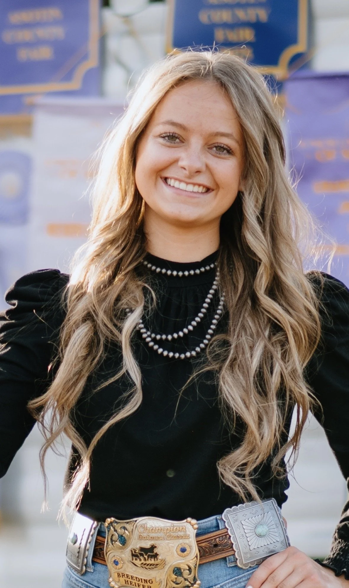 A young woman, smiling at the camera, with a black shirt on and layered jewlery..