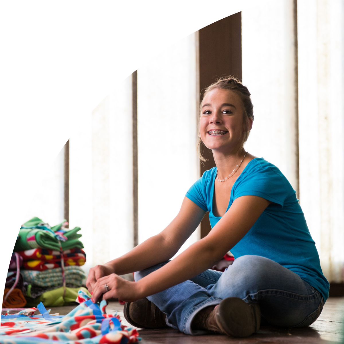 A girl with braces smiling and sitting on the floor surrounded by colorful folded fabric or clothing.