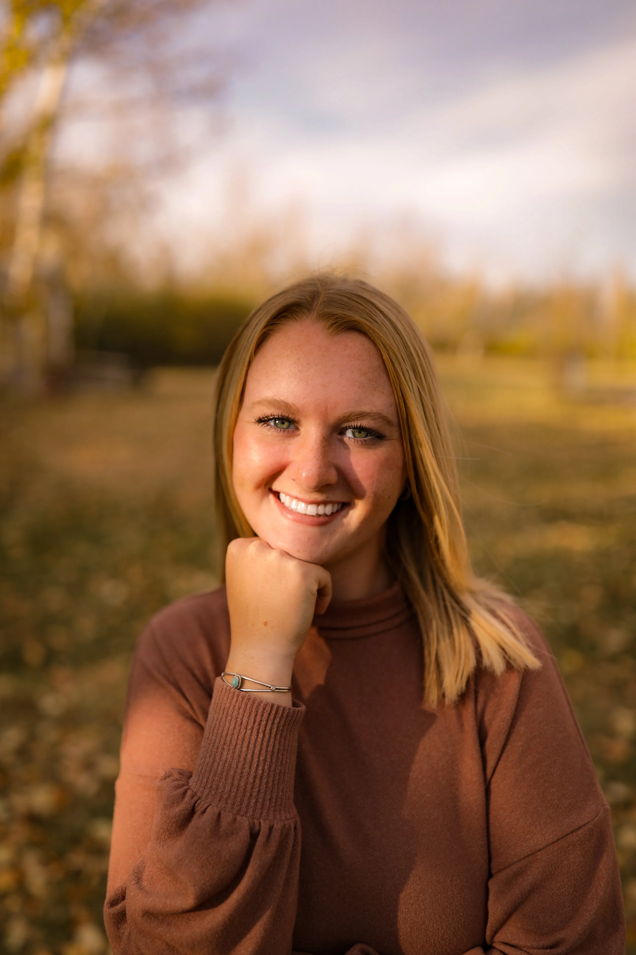A young woman with blonde hair smiling outdoors in fall, resting her chin on her hand, with a blurred background of trees and fallen leaves.