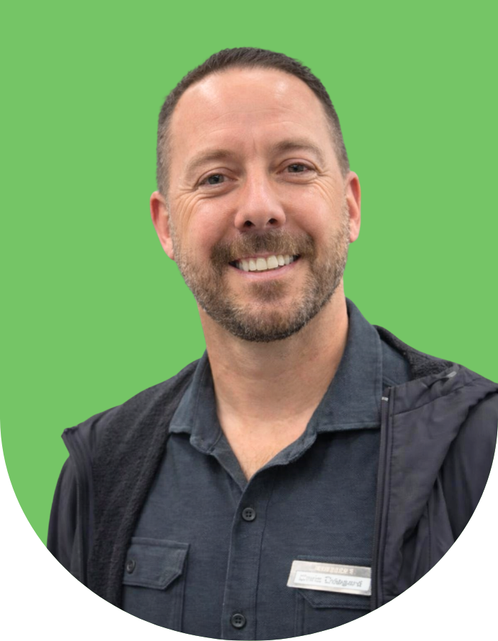 A man with short brown hair and a beard smiling at the camera, wearing a dark collared shirt and a dark jacket, against a green background.