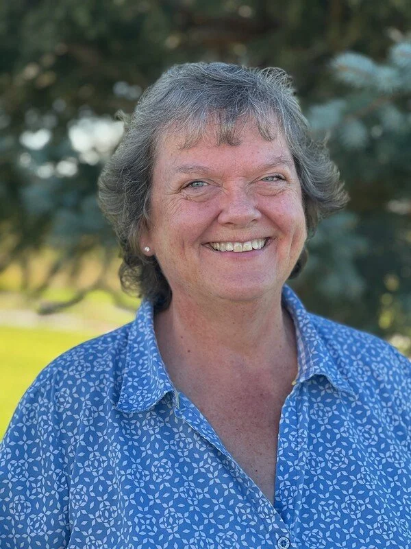 A middle-aged woman with short gray hair smiling outdoors, wearing a blue patterned button-up shirt, with trees in the background.