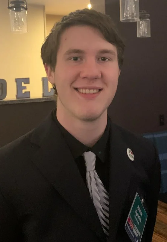 A young man with brown hair smiling in a dark suit with a pin on his lapel, wearing a striped tie, indoors with hanging light fixtures and letters in the background.