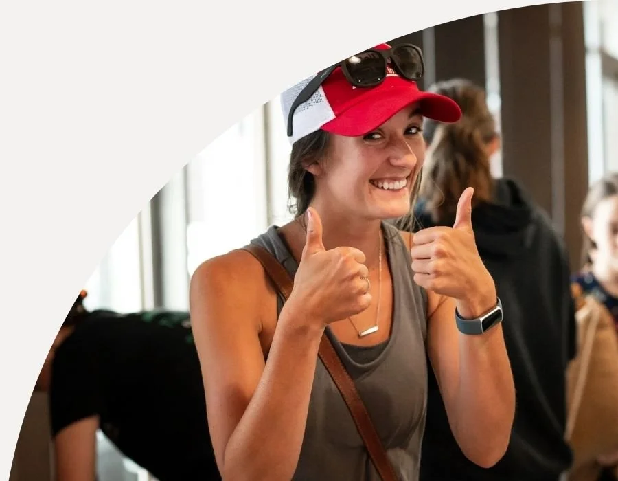 Woman smiling and giving thumbs up at indoor event, wearing a red cap with sunglasses on top, gray tank top, and a smartwatch.
