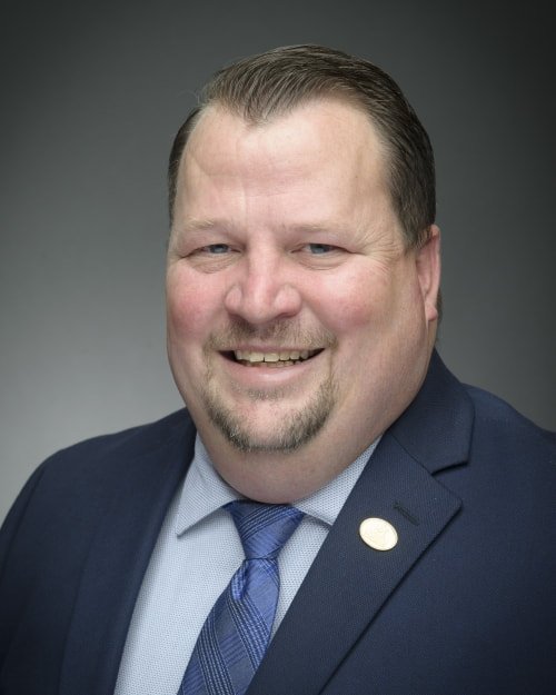 Headshot of a smiling man in a navy suit, light blue shirt, and blue tie, against a dark background.